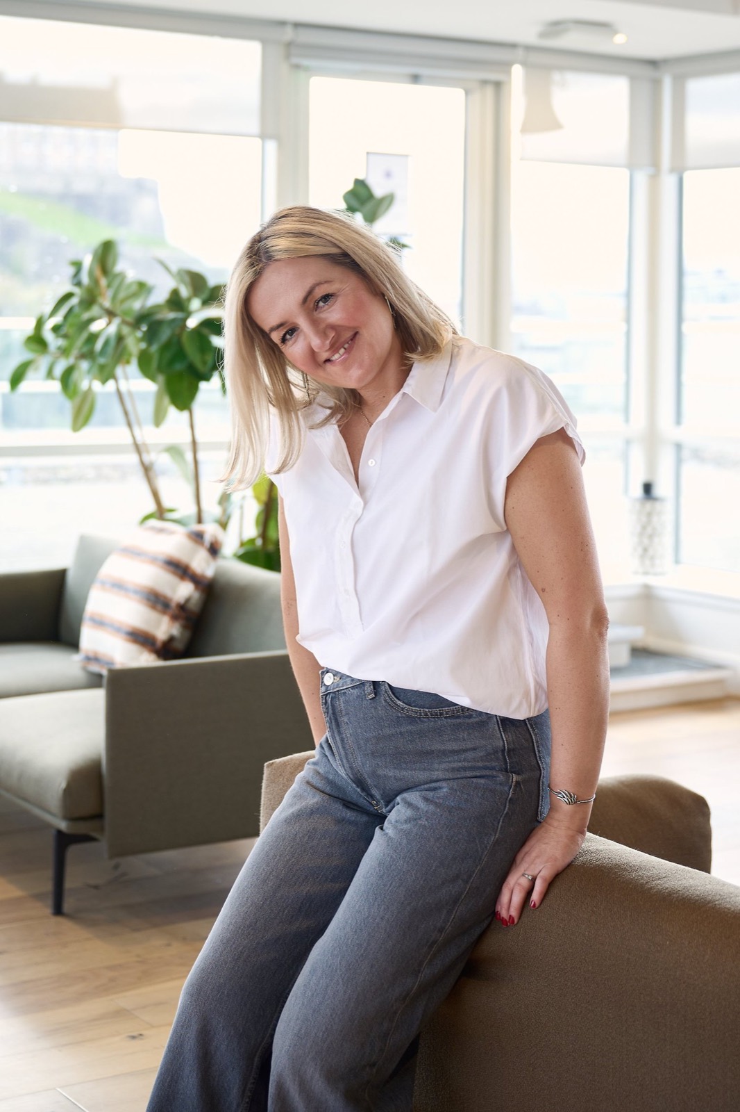 A smiling woman in a white shirt perched on a sofa arm