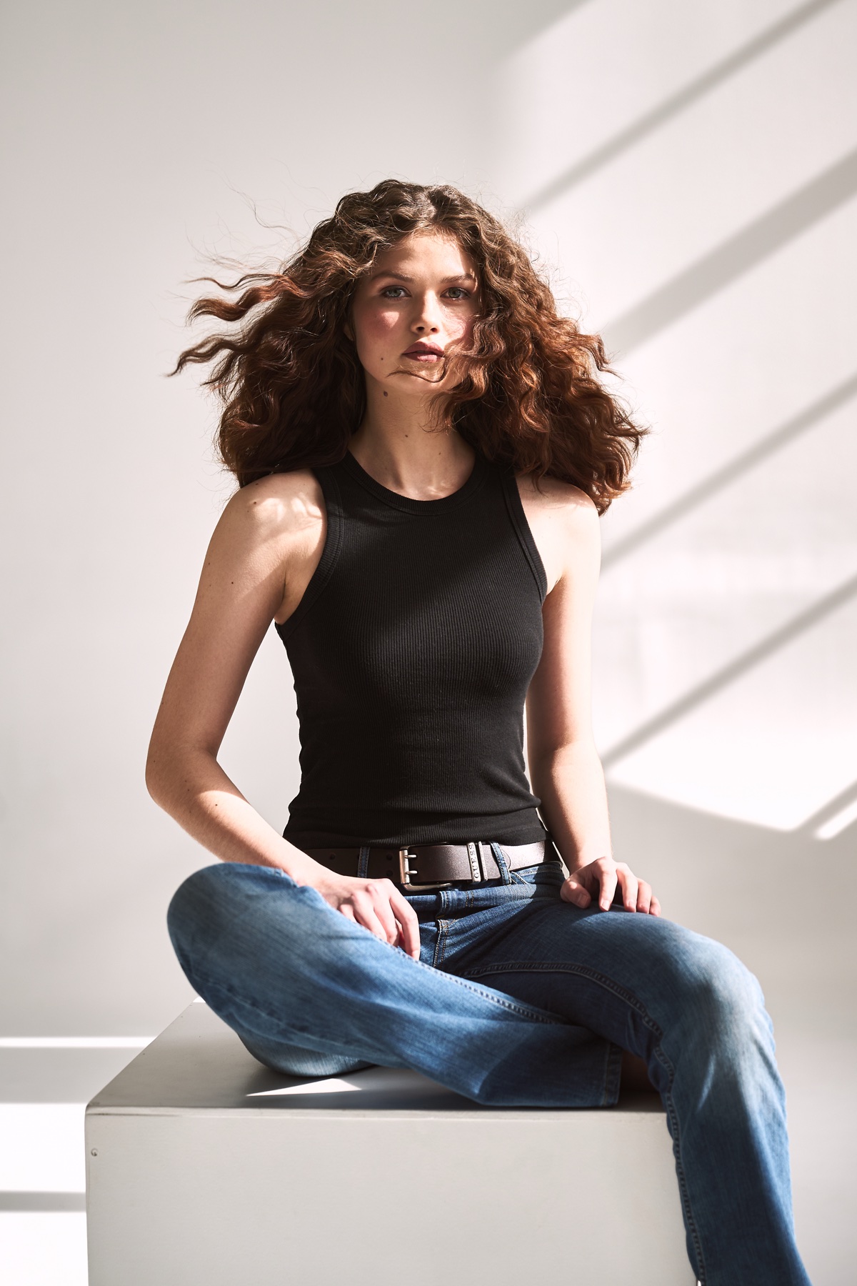 A woman with curly hair in a black top and jeans, sitting in sunlight