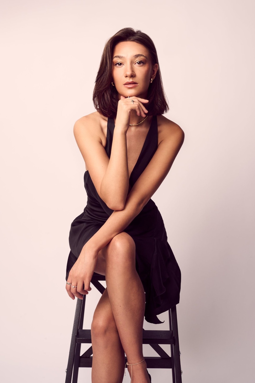 A woman in a black dress seated on a stool against blush